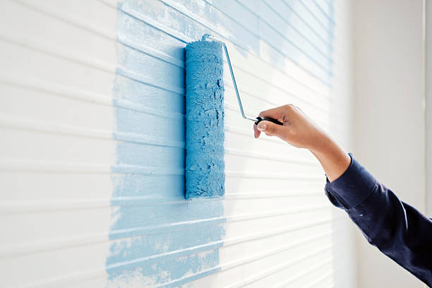 istock Close-up of a woman's hand rolling light blue paint over a white wall during a DIY home renovation. The bright, modern interior and natural light emphasize a calm, creative workspace. 2211565008