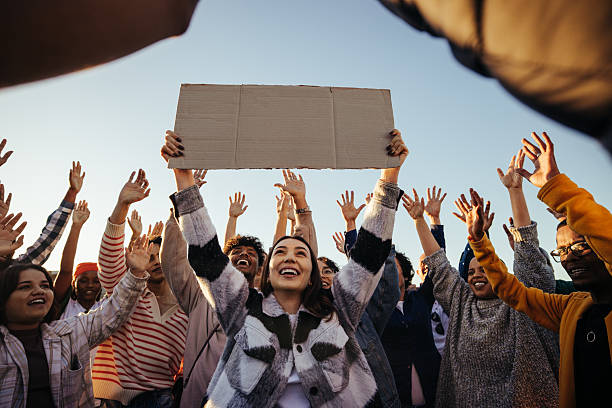 A diverse group of people gather outdoors in support of a cause, lifting their hands with enthusiasm as a centered young woman holds aloft a blank sign, exuding unity and determination.