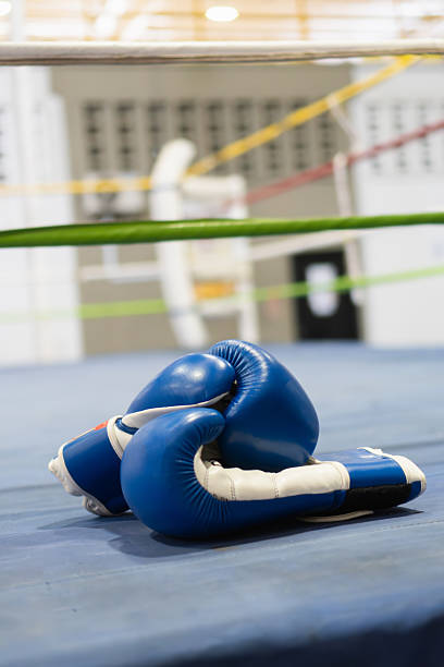 Blue boxing gloves are lying on the blue canvas of a boxing ring, ropes in the background