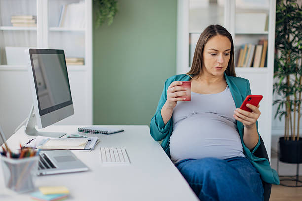 istock Pregnant business woman using mobile phone at work while sitting at desk and drinking tea 2211572998