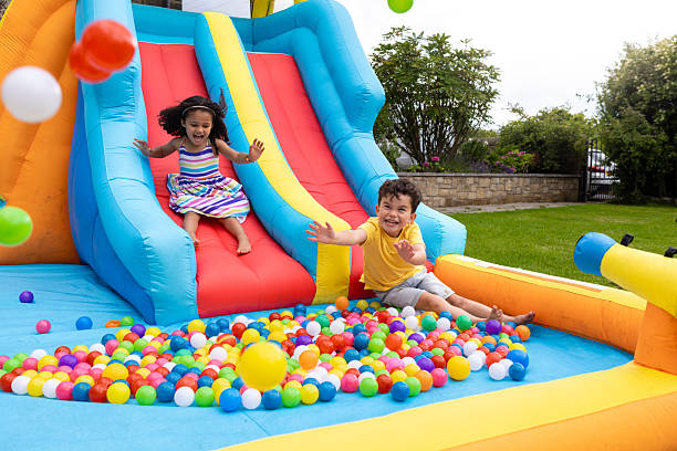 Young children playing on an inflatable slide laughing together as they go down into a ball pit. They are enjoying a day in their garden in summer in the North East of England. There are balls flying in the air around them as the boy holds his arms out to catch them.