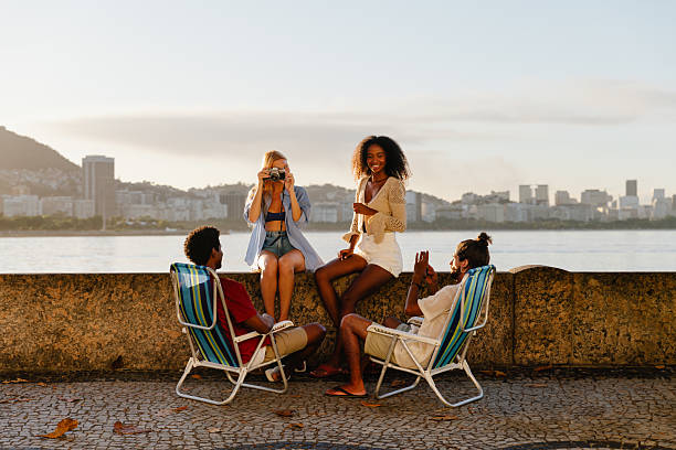 A group of young friends enjoying a relaxing evening by the waterfront at sunset. The diverse group is seated and laughing, capturing moments on camera with a city skyline as their backdrop.