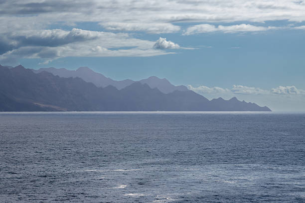 Dark volcanic coastal mountains in a fog. Calm Atlantic ocean. Blue sky with white clouds. Galdar, Las Palmas, Gran Canaria, Canary Islands, Spain.