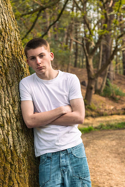 Teenage boy wearing a white t-shirt and blue denim, leaning on a tree, near a canal in a public park