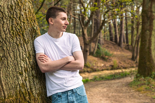 Teenage boy wearing a white t-shirt and blue denim, leaning on a tree, near a canal in a public park