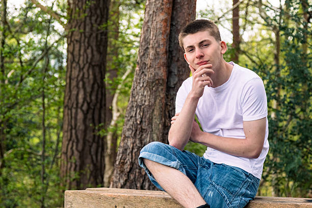 Thinking Teenage boy sititng on a bench in a public park, wearing a white t-shirt and blue denim