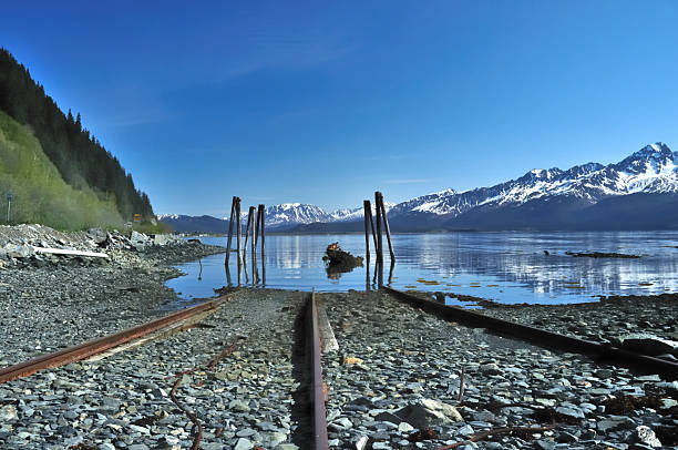 An abandoned boat ramp leads into Resurrection Bay with the Chugach Mountains in the background. An old marine motor lies in the water beneath pilings from a long gone pier.