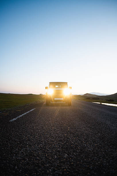 SUV with headlights on driving toward camera at sunset on an empty rural road, with mountains and grass fields in the distance.