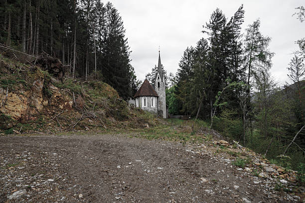 Bad Illstern, italy., Lost place. A stone church with a steeple sits nestled among tall trees, viewed from a dirt path under an overcast sky.