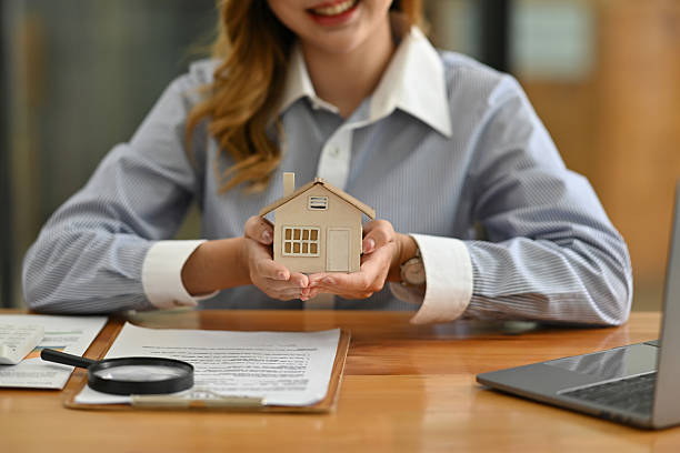 istock Close up image of Hands presenting a small house model alongside legal documents, magnifying glass, and laptop, symbolizing property sales, rental options, or quality inspections 2211632131