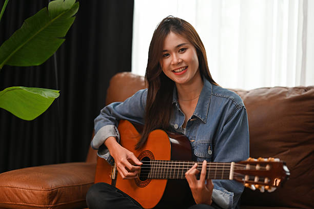 istock A young woman sits on a couch, smiling warmly while playing her acoustic guitar, radiating happiness in a cozy, natural-lit room 2211634700