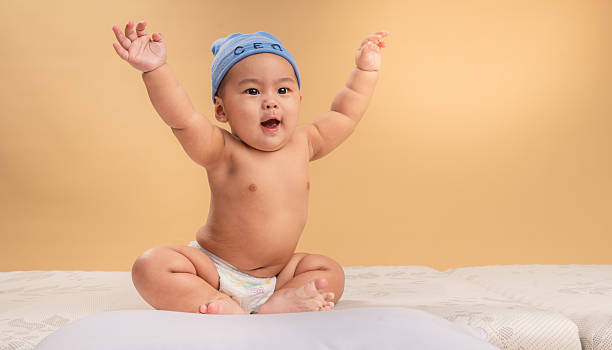 istock Asian baby wearing a blue CEO beanie sits confidently on a mattress with a curious expression. The child is shirtless, wearing a diaper, against a warm beige studio background 2211636676