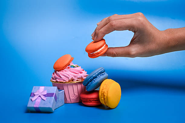 istock Hand placing orange macaron beside a pink cupcake topped with sprinkles, surrounded by colorful macarons and a blue gift box on a pastel blue background 2211638357