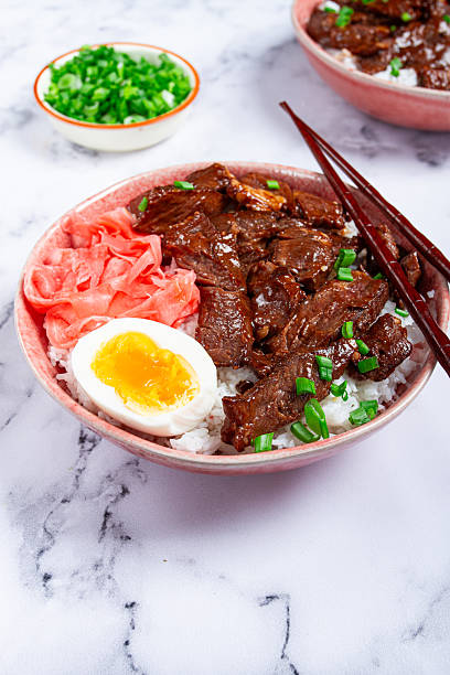 Gyudon Japanese beef with rice, pickled ginger, boiled egg slice, garnished with green onions, in a bowl with chopsticks,