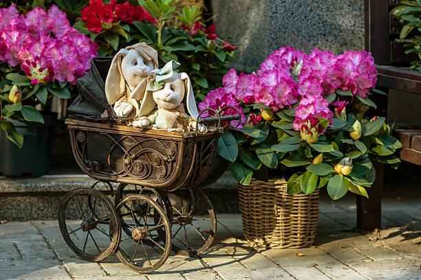 Two plush bunnies in an antique pram framed by vibrant pink rhododendrons create a warm vintage scene that evokes spring, Easter, childhood nostalgia and romantic charm.