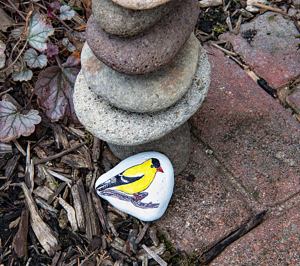 Painted rock of American Goldfinch placed by stack of rocks in nature setting.