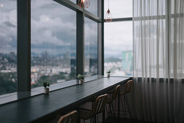 Contemporary co-sharing office space featuring a panoramic cityscape view through large floor-to-ceiling windows. The interior includes a long communal work counter with bar stools, minimalist hanging pendant lights, and small potted plants for decoration. Captures the modern work environment, ideal for concepts such as flexible workspaces, remote working, business hubs, and urban office culture