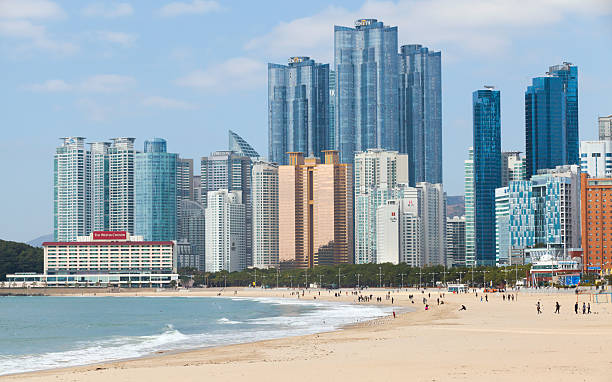 Busan, South Korea - March 17, 2018: Skyline of tall, modern buildings bordering a sandy beach, with people strolling and calm waters embracing the shore under a clear sky. Haeundae beach landscape