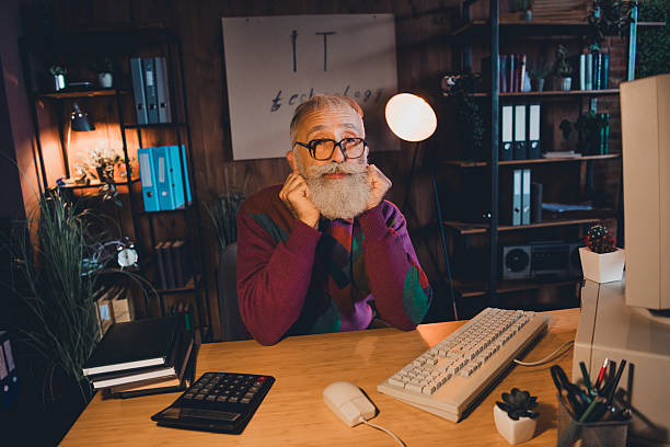 istock Elderly programmer working at desk in casual sweater in cozy home office setting under warm evening light 2211717781