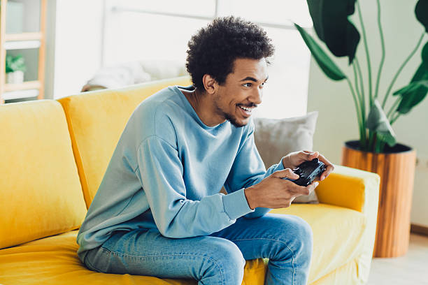 istock Young man enjoying indoor leisure time with a video game controller, smiling on a yellow couch in a bright modern home interior. 2211718087