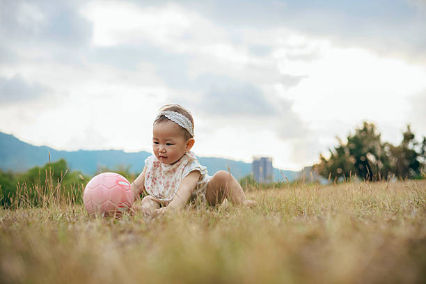 An Asian baby girl plays with a ball on the grass, capturing a pure moment of exploration and growth in a natural setting. The scene also highlights society's emphasis on child welfare, public education, and healthy development, reflecting how modern families balance economic pressures with policy support to nurture early childhood growth and parent-child bonding.