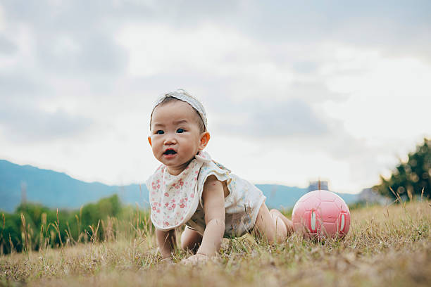 An Asian baby girl plays with a ball on the grass, capturing a pure moment of exploration and growth in a natural setting. The scene also highlights society's emphasis on child welfare, public education, and healthy development, reflecting how modern families balance economic pressures with policy support to nurture early childhood growth and parent-child bonding.