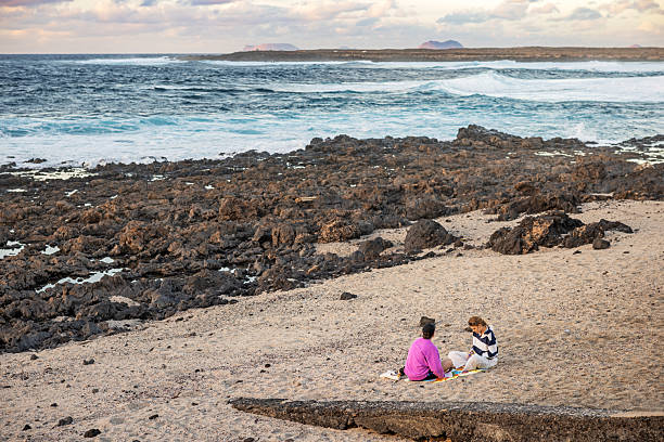 La Santa, Lanzarote, Canary Islands, Spain - March 21th 2025:  This stock photo captures a serene moment of two women sitting on a sandy beach surrounded by rugged rocks, as they wait for the sunset. The women are seated on a colorful blanket, engaged in conversation, with the vast ocean and distant islands in the background. The waves crash gently against the rocky shore, creating a peaceful and picturesque scene. The sky is adorned with soft clouds, adding to the tranquil ambiance of the setting. This image is interesting and relevant as it portrays a calm and reflective moment in nature, highlighting the beauty of the coastal landscape and the simple joy of spending time by the sea.