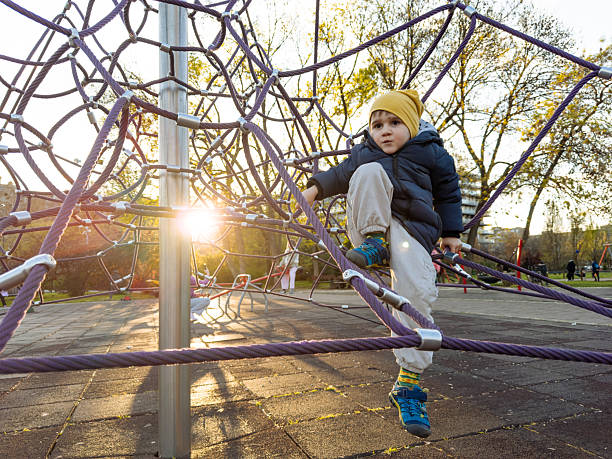 Smiling toddler resting on a playground rope net. Child enjoys playful outdoor activity while developing confidence, balance and motor skills in an autumn park.