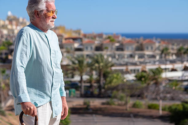 istock Senior caucasian man with back problems walking with the help of a cane. Elderly people outdoor face the sea, horizon over water 2211759546