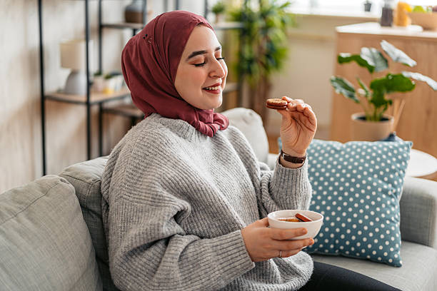 istock Young Muslim Woman Eating Cookies While Sitting On The Sofa 2211759710