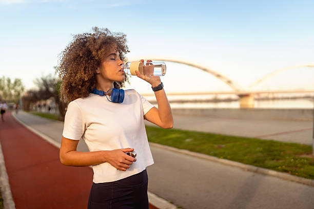 istock Fit young woman hydrating after a workout while standing near a riverfront path with a bridge in the background, wearing sporty attire and headphones. 2211768337