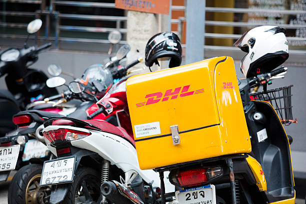 Back of German DHL express delivery box in motorcycle. Motorcycle is parked among other motorcycles in small street and Soi of Phloen Chit Road near Phloen Chit BTS station. On box is banner for sustainable development goals 2050 with net zero emissions
