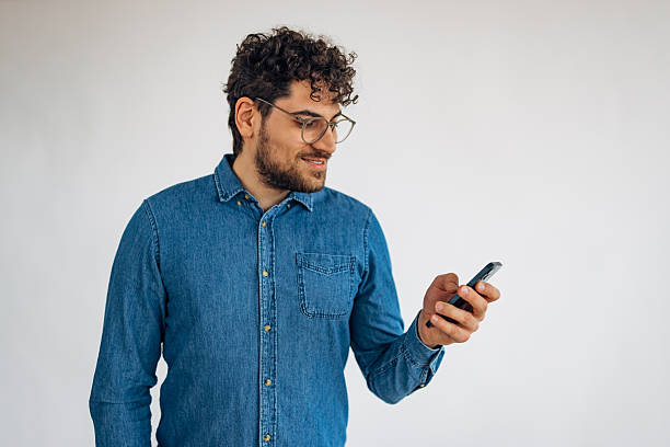 istock Smiling man in eyeglasses typing SMS on white background using mobile phone 2211793127