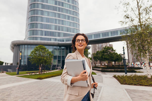 istock Businesswoman walking outside office building carrying laptop and smiling 2211808564