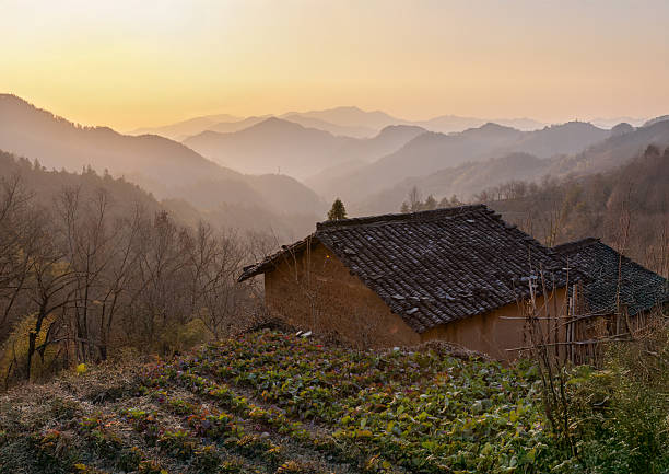 At sunset, layers of misty mountains stretch into the distance as an earthen house stands quietly beside a terraced field. The golden sky and soft light evoke a sense of stillness and timeless rural beauty in the heart of Anhui.
