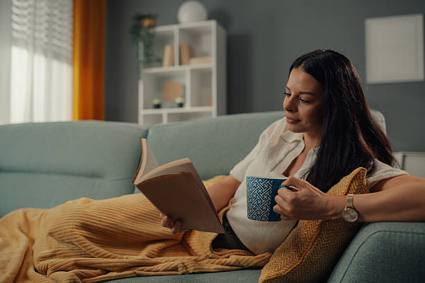 istock Young woman relaxing on sofa reading a book and drinking tea 2211835753