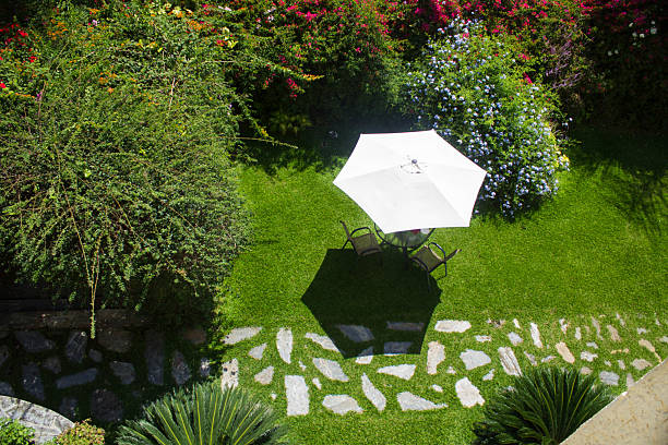High-angle view of a serene garden setting. A white parasol shades a small table and chairs on lush green grass, surrounded by vibrant flowering bushes and a stone pathway. Perfect for illustrating relaxation, outdoor living, and peaceful escapes.