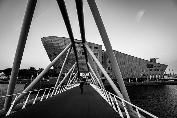 Amsterdam, Netherlands - July 3, 2018: This black-and-white photograph captures the NEMO Science Museum in Amsterdam, Netherlands, from a pedestrian bridge leading to the building. The composition frames a solitary pedestrian walking towards the entrance.