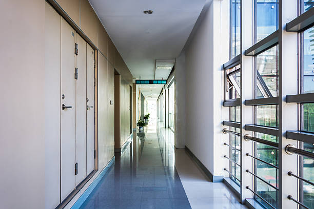 A cleanly lit hospital hallway with a polished reflective floor, visible emergency signs, and essential equipment like fire extinguishers. The corridor's perspective leads to multiple rooms with doors ajar and a welcoming atmosphere, with natural light enhancing its clinical yet friendly design. Ideal for representing healthcare and professional environments.