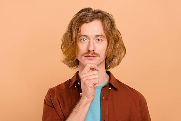 Portrait of attractive content bewildered red-haired guy touching chin making decision isolated over beige pastel color background.