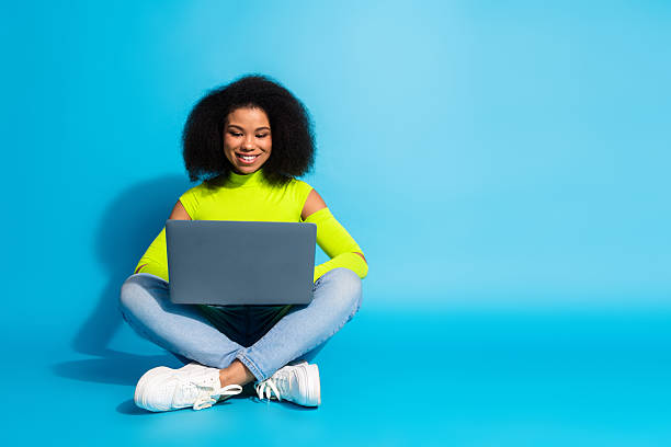 istock Young woman sitting cross-legged using a laptop against a blue backdrop, wearing a neon green top and smiling brightly. 2211889748