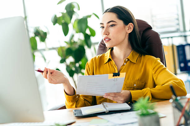 Qualified female professional reviewing documents while working in a modern office environment Businesswoman analyzing paperwork at her desk in a brightly lit modern office, exemplifying focus, professionalism, and a productive working atmosphere. qualified report stock pictures, royalty-free photos & images