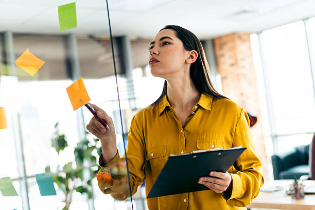 istock Qualified businesswoman reviewing post-it notes on a glass board in a modern office setting, showcasing creativity and organization. 2211890030
