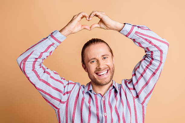istock Cheerful young man making a heart gesture with his hands while smiling in a casual striped shirt on a beige background. 2211890422