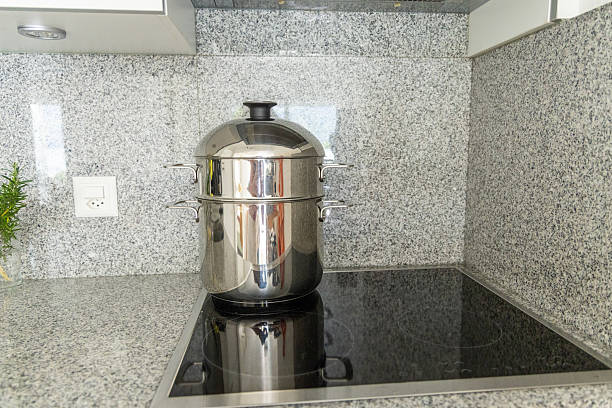Shiny stainless steel steamer pot sits on an induction stove in a granite-topped kitchen. Modern cooking appliance for healthy meals. Contemporary kitchen interior.