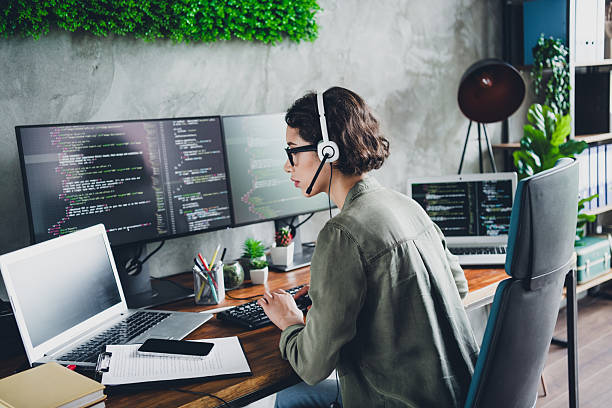 istock Young female programmer working at office desk with multiple screens, wearing casual attire, focused on coding and programming 2211894130