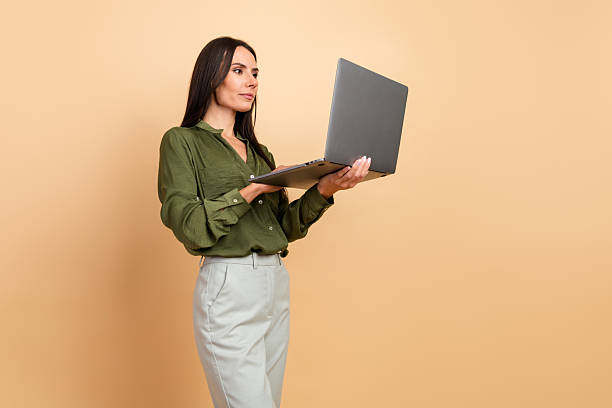 istock Professional woman working on a laptop while standing against a beige background in formal attire and showcasing confidence 2211894750