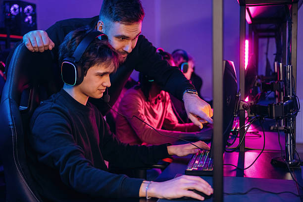 istock Guy is standing and looking, helping. Gamers are playing video game by computers 2211900639
