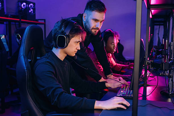 istock Guy is standing and looking, helping. Gamers are playing video game by computers 2211900654