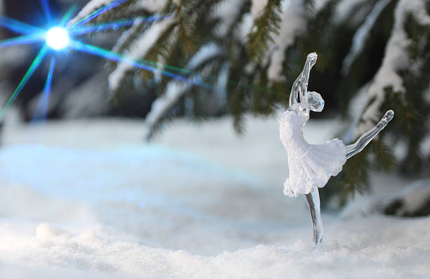 A glass or crystal figurine of a ballerina stands on a snow-covered ground, poised in an elegant arabesque. The translucent figure, dressed in a delicately detailed tutu, catches the light, adding a shimmering effect. Snow-draped evergreen branches form the background, enhancing the wintry atmosphere. A bright, star-like flare of light glows in the upper left corner, adding a mystical element to the serene scene.